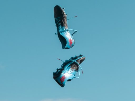 pair of blue-and-black New Balance cleats in the air under teal sky with white clouds